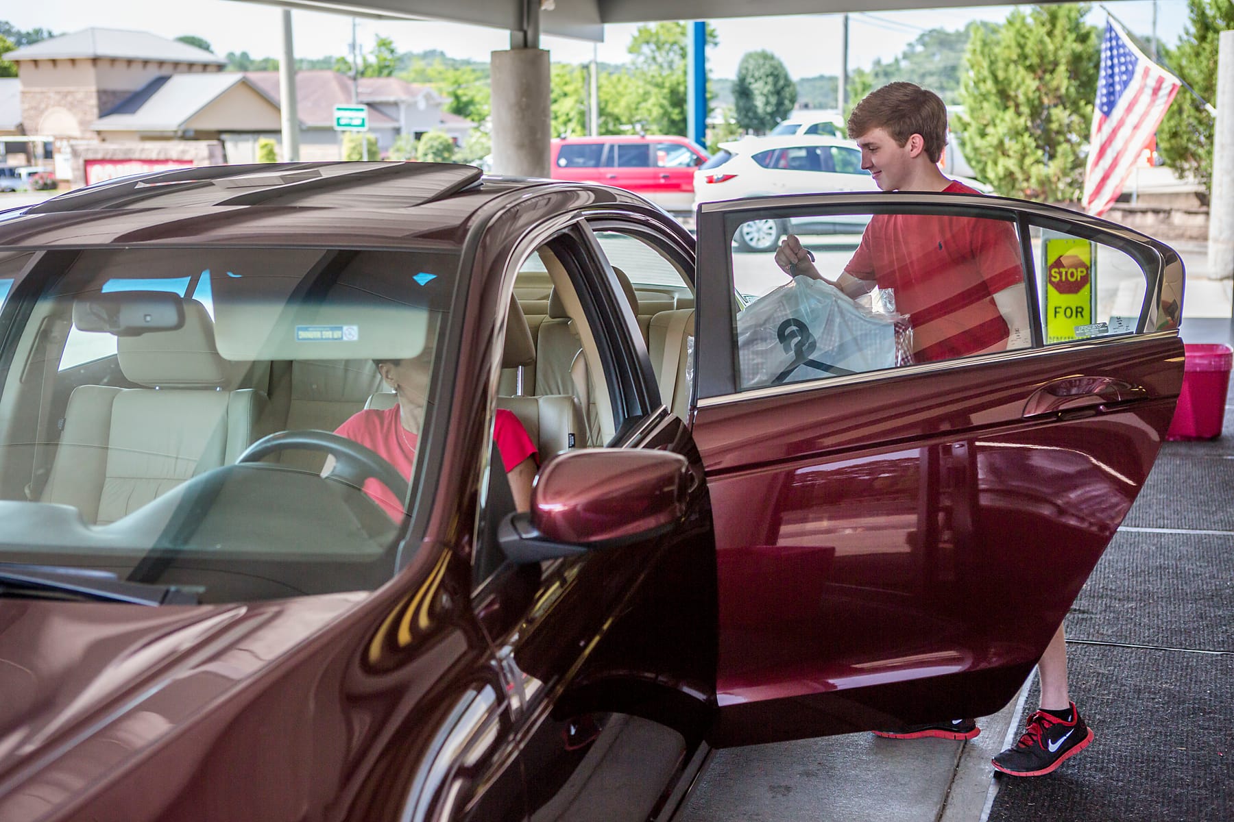 A Complete Cleaners team member in a red polo handing finished, hung garments to a customer at the drive-thru pickup window